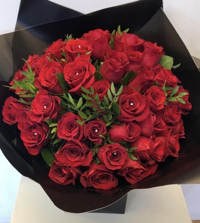 Luxury hand-tied bouquet of 50 large-headed red Freedom roses with sparkling diamanté centres and complementary foliage, wrapped in luxury cellophane and presented in a signature black gift bag with rope handles.