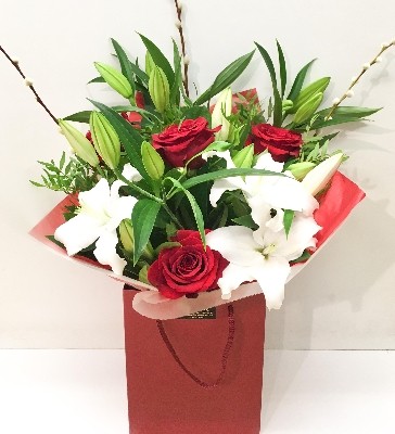 Hand-tied bouquet featuring luxury red Freedom roses, white lilies, and willow accents, wrapped in luxury cellophane and matching gift bag, hand-tied in water.