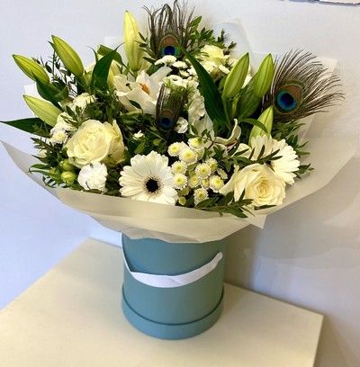 Elegant white bouquet featuring lilies, roses, alstroemeria, and chrysanthemums, accented with vibrant peacock feathers and presented in a luxury hatbox.