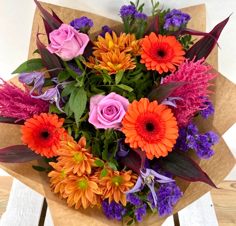 Vivid mix of orange, pink and purple tones, design includes roses, astilbe and clematis along with other summer florals hand tied in water. 