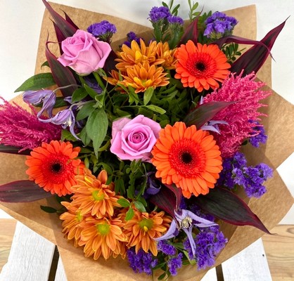 Vivid mix of orange, pink and purple tones, design includes roses, astilbe and clematis along with other summer florals hand tied in water. 