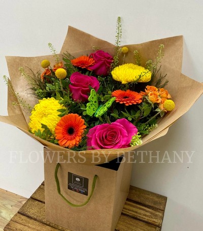 A bright bouquet in orange, yellow and cerise. Including roses, germini, spray rose, green bell and more. Presented in a kraft bag and wrap. 