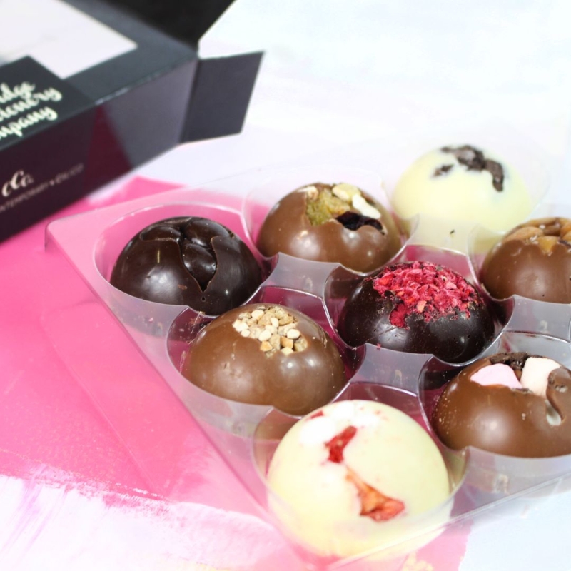 Assorted luxury solid chocolate domes from The Cambridge Confectionery Co., featuring flavours like Dark Raspberry, Milk Honeycomb, Dark Salted Caramel, and more, arranged in gift boxes.