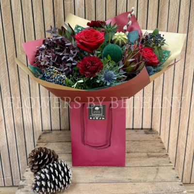Luxury red Christmas bouquet with deep festive tones, winter textures and seasonal greenery arranged in a gift bag.