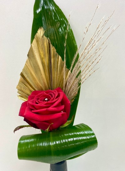  Single red rose displayed in a slim black ceramic vase, accented with gold spear, baby pampas grass, and folded aspidistra leaves.