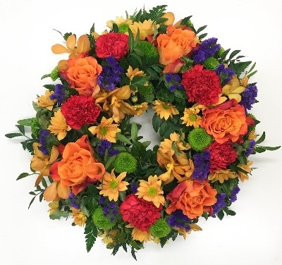 Sunset-themed funeral wreath with orange, red, and purple flowers, accented with green filler florals on a lush foliage base.