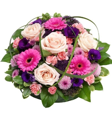 Pink and purple funeral posy pad with mixed seasonal flowers, complementary foliage, and a textured grass caging effect.