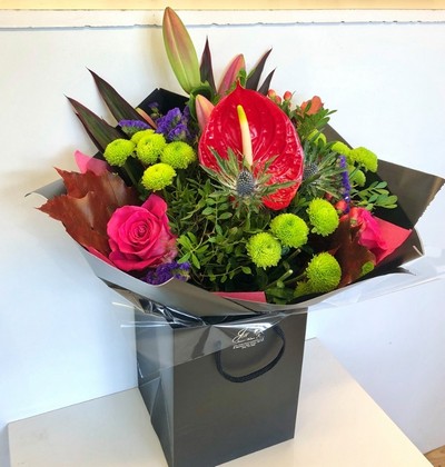 A beautiful bouquet including pink roses and lily, red anthurium and berries, green kermit chrysanthemum and pop's or purple statice and blue thistle all wrapped in black cellophane and presented in a gift bag. 