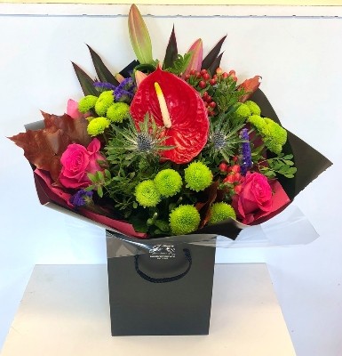 A beautiful bouquet including pink roses and lily, red anthurium and berries, green kermit chrysanthemum and pop's or purple statice and blue thistle all wrapped in black cellophane and presented in a gift bag. 