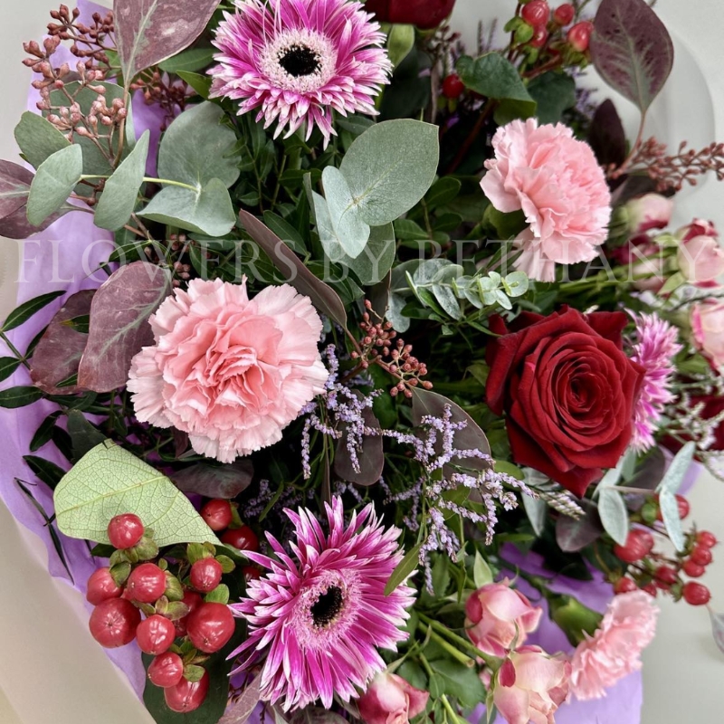 Berry Christmas bouquet featuring red roses, pink germini, carnations, spray roses, lilac limonium, and hypericum berries, wrapped in cellophane and presented in a burgundy gift bag.
