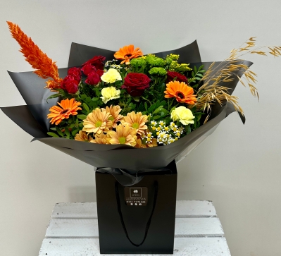 A bouquet containing roses, spray rose, germini, matricaria, chrysanthemum, carnations, solidago, celosia and corn finished with black cellophane and placed in a black bag.