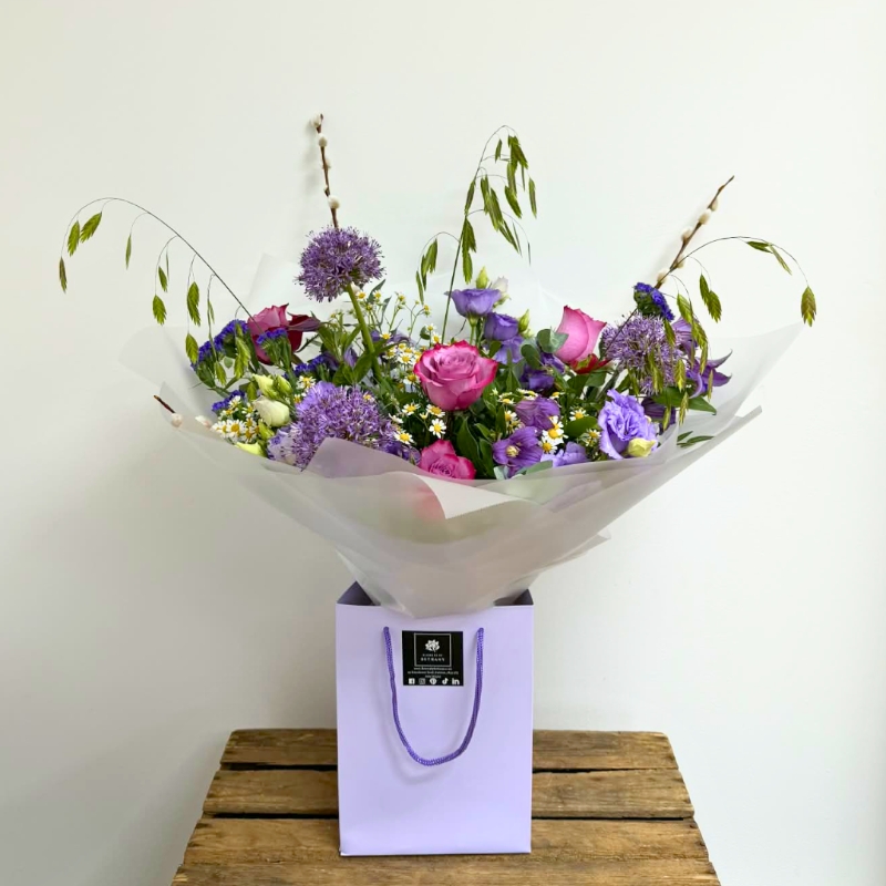 Lilac garden-inspired bouquet featuring seasonal lilac and purple florals with soft texture and natural movement, hand-tied with mixed foliage in a luxury lavender gift bag by Flowers by Bethany.
