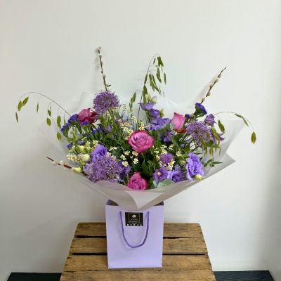 Lilac garden-inspired bouquet featuring seasonal lilac and purple florals with soft texture and natural movement, hand-tied with mixed foliage in a luxury lavender gift bag by Flowers by Bethany.