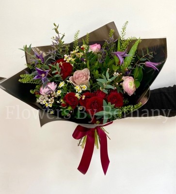 Hand-tied bouquet featuring ranunculus, roses, matricaria, clematis, and seasonal florals, presented in signature black wrap with a satin bow.
