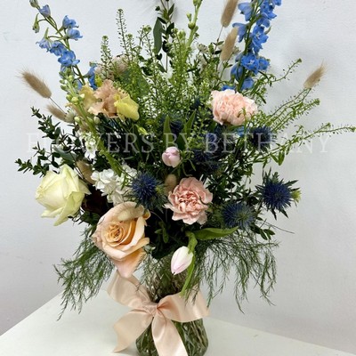 A hand tied bouquet filled with seasonal varities in peach, pink and blue tones presented in a glass vase with a bow. 