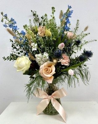 A hand tied bouquet filled with seasonal varities in peach, pink and blue tones presented in a glass vase with a bow. 