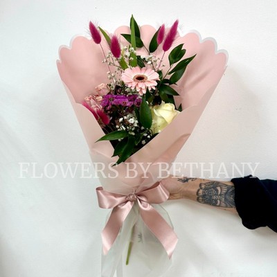 A petite hand tied bouquet in mixed pink hues in a pink scalloped wrap with positivity script. 