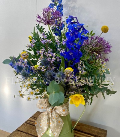 A sage zinc jug filled with wild meadow florals including delphinium, allium, daffodils, tulips and more, finished with a matching bow. 