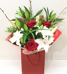 Hand-tied bouquet featuring luxury red Freedom roses, white lilies, and willow accents, wrapped in luxury cellophane and matching gift bag, hand-tied in water.