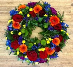Elegant funeral wreath with vibrant flowers in orange, red, blue, purple, green, and yellow, accented with lush foliage.