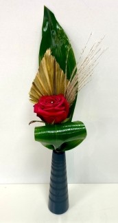  Single red rose displayed in a slim black ceramic vase, accented with gold spear, baby pampas grass, and folded aspidistra leaves.