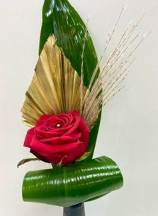  Single red rose displayed in a slim black ceramic vase, accented with gold spear, baby pampas grass, and folded aspidistra leaves.