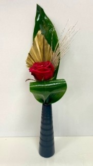  Single red rose displayed in a slim black ceramic vase, accented with gold spear, baby pampas grass, and folded aspidistra leaves.