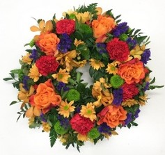 Sunset-themed funeral wreath with orange, red, and purple flowers, accented with green filler florals on a lush foliage base.