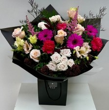 A bouquet containing roses, spray roses, germini, lizianthus, carnations, limonium, astilbe and skimmia finished in black cellophane and placed in a black gift bag.