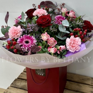 Berry Christmas bouquet featuring red roses, pink germini, carnations, spray roses, lilac limonium, and hypericum berries, wrapped in cellophane and presented in a burgundy gift bag.