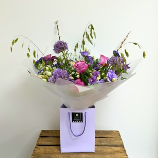 Lilac garden-inspired bouquet featuring seasonal lilac and purple florals with soft texture and natural movement, hand-tied with mixed foliage in a luxury lavender gift bag by Flowers by Bethany.