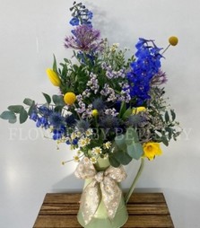 A sage zinc jug filled with wild meadow florals including delphinium, allium, daffodils, tulips and more, finished with a matching bow. 