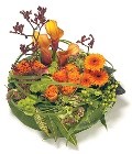 Modern funeral posy with orange, gold, and green seasonal flowers, wrapped in grasses, leaves, and mixed foliage for a contemporary look.
