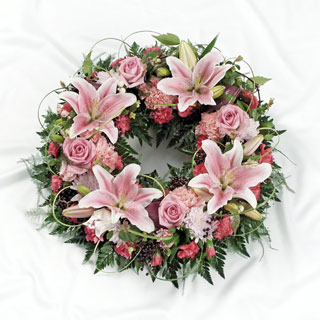 Soft pink funeral wreath with lilies, roses, chrysanthemums, and carnations, accented with foliage and grass loops.