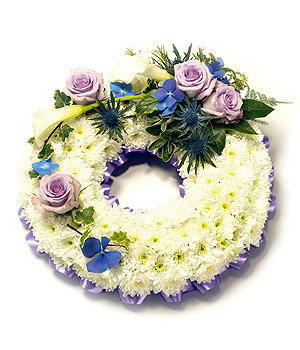 White chrysanthemum funeral wreath with a lilac and blue floral focal, accented with complementary flowers and mixed foliage.